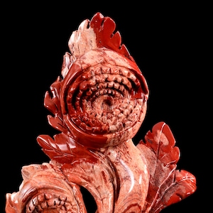 May include: A polished red and cream-coloured stone carving depicting a stylised plant with a large, circular, tooth-like central opening and multiple leaf-shaped protrusions extending from the base and sides of the main structure.