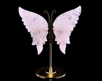 May include: A decorative butterfly sculpture with translucent, pale pink wings. The wings are supported by a gold-colored metal stand with a circular base. The butterfly's body is also gold-colored, and the background is black.