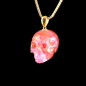 May include: A gold chain necklace with a skull pendant. The skull is carved and displays pink and orange tones. The eye sockets and nose cavity are clearly visible. The background is black.