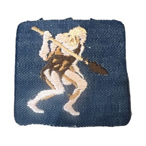 May include: A blue denim patch with an embroidered image of a person holding a spear. The person is depicted in a light brown and pink color scheme.