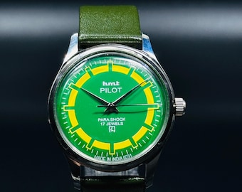 May include: A vintage HMT Pilot watch with a green and yellow dial and a green leather strap. The watch face features the text "PILOT" and "PARA SHOCK 17 JEWELS". The watch has a silver-colored case and is marked "MADE IN INDIA 0231".