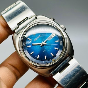 May include: A vintage Seiko wristwatch with a blue dial and a silver-tone metal strap. The watch face shows the day and date, with the word "SEIKO" visible. The watch has a rectangular case and a brushed metal finish.