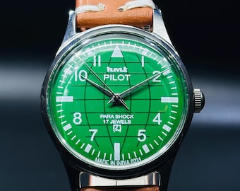 May include: A green analog wristwatch with a brown leather band. The watch face has a white dial with black markings and the words "HMT PILOT PARA SHOCK 17 JEWELS" printed on it. The watch is made in India.