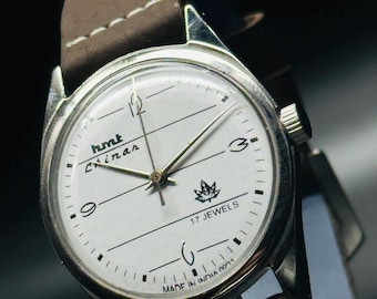 May include: A silver wristwatch with a white face and brown leather band. The watch face has black markings and the text "hmt Chinar 17 JEWELS MADE IN INDIA 0031".