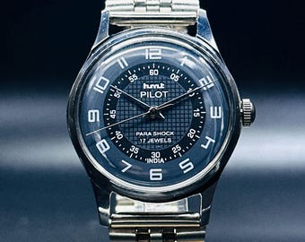 May include: A silver wristwatch with a black face and silver hands. The watch face has white markings for the hours and minutes, and the words "humt PILOT PARA SHOCK 17 JEWELS INDIA" are printed on the face. The watch has a silver metal band.