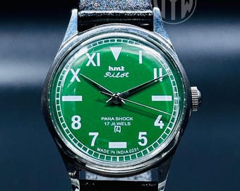 May include: A green-faced wristwatch with silver accents and a brown leather band. The watch face has Roman numeral markings and the text "hmt Pilot" and "PARA SHOCK 17 JEWELS" in white. The watch also has the text "MADE IN INDIA 0231" printed on the face.