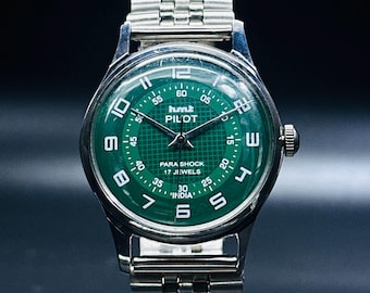 May include: A silver wristwatch with a green face. The face has white markings and the text "humt PILOT PARA SHOCK 17 JEWELS INDIA".