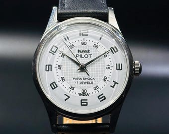 May include: A silver wristwatch with a white face and black leather band. The watch face has black Roman numeral hour markers, black hands, and a small seconds dial. The watch face also has the text "hmt PILOT PARA SHOCK 17 JEWELS 30 'INDIA'".