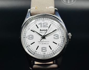 May include: A white analog wristwatch with a silver case and a brown leather band. The watch face has black hands and black markers for the hours. The watch face also has the text "hmt PILOT PARA SHOCK 17 JEWELS".