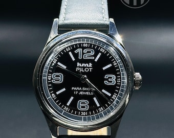 May include: A black-faced wristwatch with silver accents and a brown leather band. The watch face has white markings and the text "hmt PILOT PARA SHOCK 17 JEWELS".