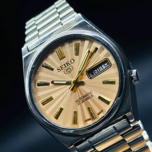 May include: A vintage Seiko 5 automatic wristwatch with a silver stainless steel strap and case. The watch face is a light tan colour with gold hour markers and hands. The day and date are displayed at the 3 o'clock position. The watch is marked "AUTOMATIC 17 JEWELS" and "JAPAN MADE".