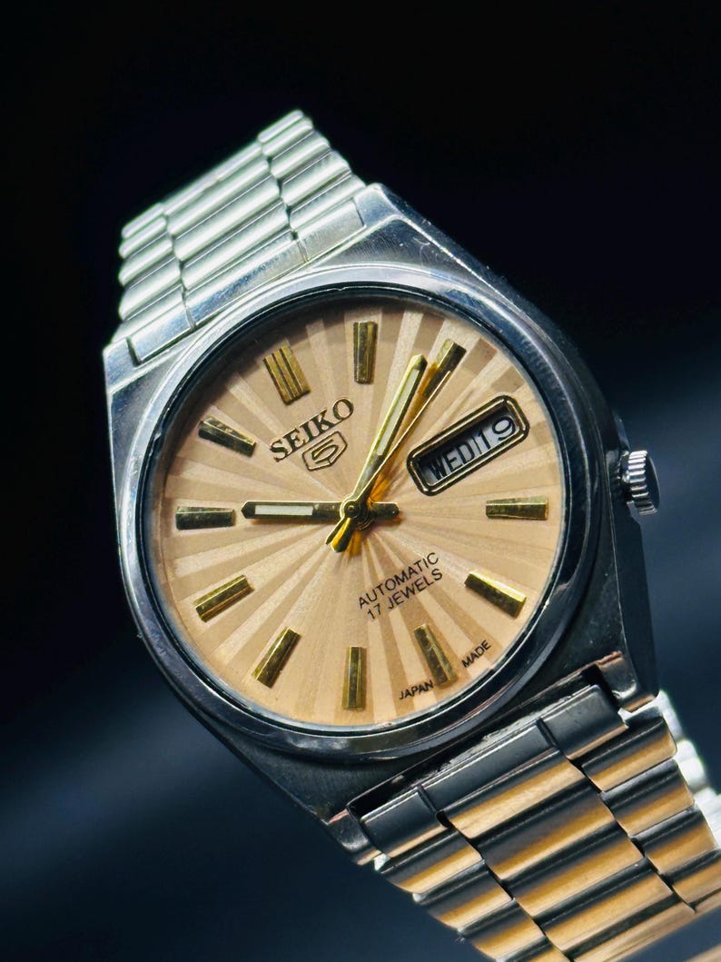 May include: A vintage Seiko 5 automatic wristwatch with a silver stainless steel strap and case. The watch face is a light tan colour with gold hour markers and hands. The day and date are displayed at the 3 o'clock position. The watch is marked "AUTOMATIC 17 JEWELS" and "JAPAN MADE".