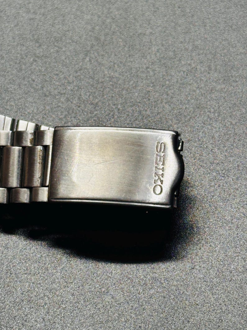 May include: A close-up shot of a silver Seiko watch clasp. The rectangular clasp features the engraved brand name "SEIKO". The watch strap is composed of numerous small, connected metal links.
