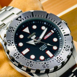 Può includere: Un orologio automatico Seiko 5 in acciaio inossidabile con quadrante nero. L'orologio presenta indici bianchi, un display giorno-data e una lunetta girevole con indicazioni dei minuti. Anche il cinturino è in acciaio inossidabile.