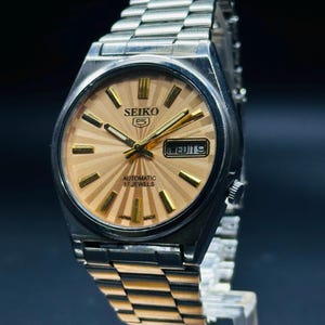 May include: A vintage Seiko 5 automatic wristwatch with a silver stainless steel strap and case. The watch face is light brown with gold markers and hands. The watch displays the day and date. The text "SEIKO 5", "AUTOMATIC 17 JEWELS", "JAPAN MADE" is visible.