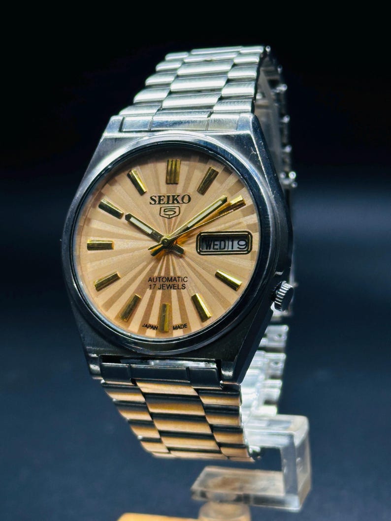 May include: A vintage Seiko 5 automatic wristwatch with a silver stainless steel strap and case. The watch face is light brown with gold markers and hands. The watch displays the day and date. The text "SEIKO 5", "AUTOMATIC 17 JEWELS", "JAPAN MADE" is visible.