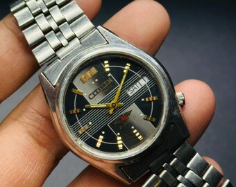 May include: A vintage Citizen automatic watch with a black dial and silver-tone metal band. The watch face features gold-tone hands and markers, with the day and date displayed. The watch is held in a hand.