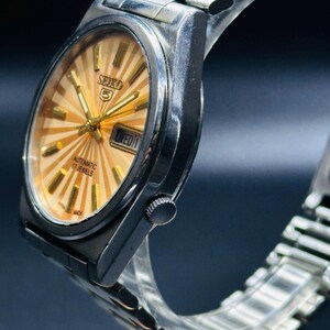 May include: A silver Seiko 5 automatic wristwatch with a gold sunburst dial. The watch has a day-date display and a silver metal strap. The watch face features gold hands and markers. The watch is marked "Japan Made".