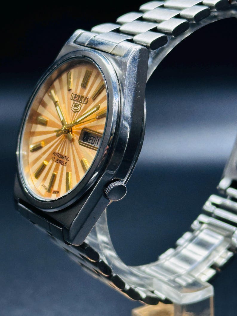 May include: A silver Seiko 5 automatic wristwatch with a gold sunburst dial. The watch has a day-date display and a silver metal strap. The watch face features gold hands and markers. The watch is marked "Japan Made".