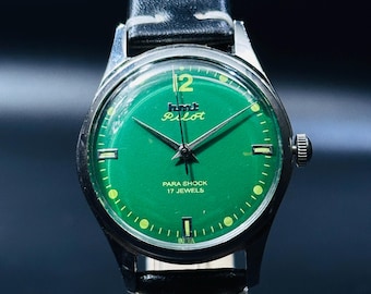 May include: A green-faced wristwatch with a black leather band. The watch face has white markings and the words "HMT Pilot" and "Para Shock 17 Jewels".