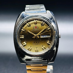 May include: A gold-toned wristwatch with a silver band. The watch face has black Roman numeral hour markers, a date window, and the text "Crystal 21 Jewels".