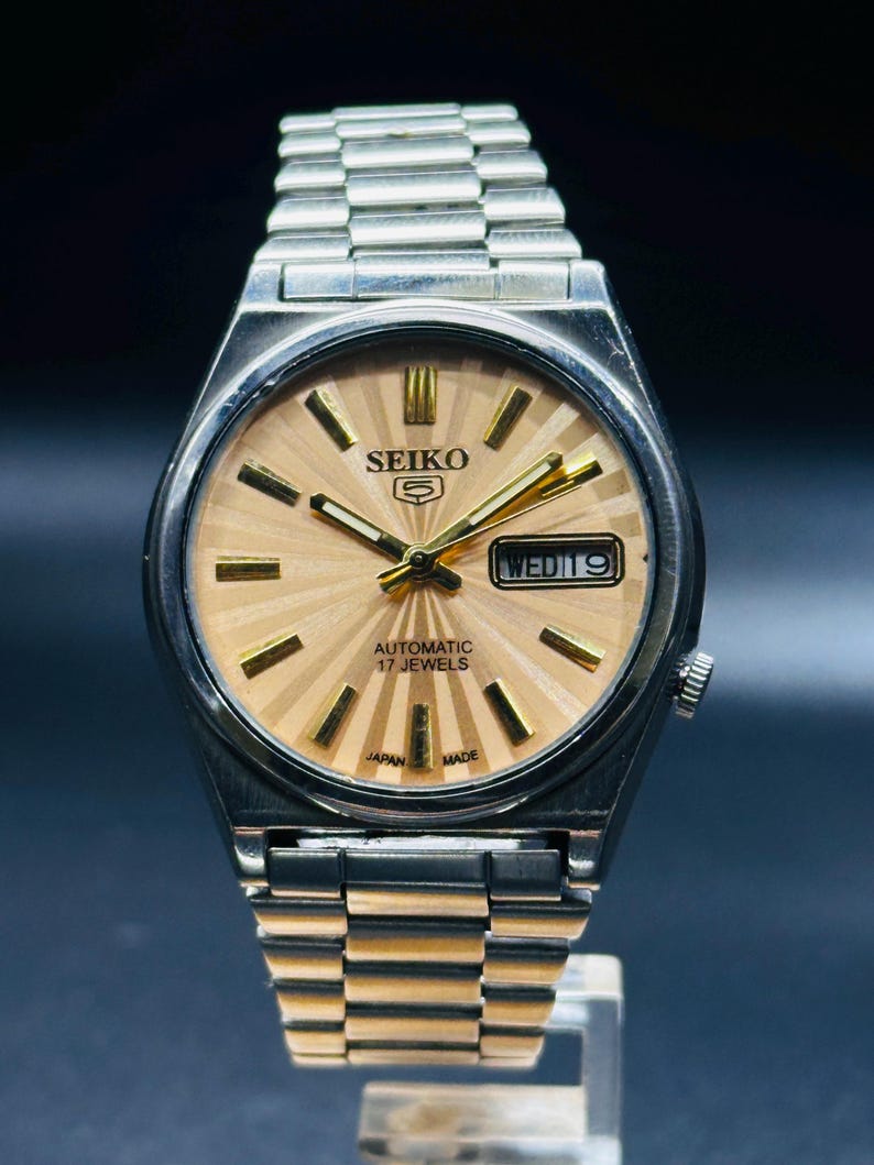May include: A vintage Seiko 5 automatic wristwatch with a silver stainless steel strap and case. The watch face is a light gold colour with gold hands and markers. The day and date are displayed at the 3 o'clock position. The watch reads "AUTOMATIC 17 JEWELS" and "JAPAN MADE".