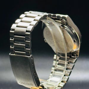 May include: A silver wristwatch with a stainless steel strap. The watch face is not visible, but the strap and clasp are in focus. The clasp is engraved with the word "SEIKO". The watch is displayed on a clear stand against a dark background.