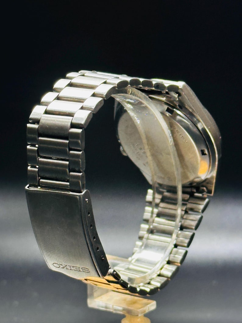 May include: A silver wristwatch with a stainless steel strap. The watch face is not visible, but the strap and clasp are in focus. The clasp is engraved with the word "SEIKO". The watch is displayed on a clear stand against a dark background.
