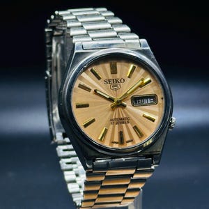 May include: A vintage Seiko automatic watch with a silver stainless steel strap and case. The watch face is light brown with gold hands and markers. The watch displays the day and date. The text "SEIKO" is visible on the watch face.