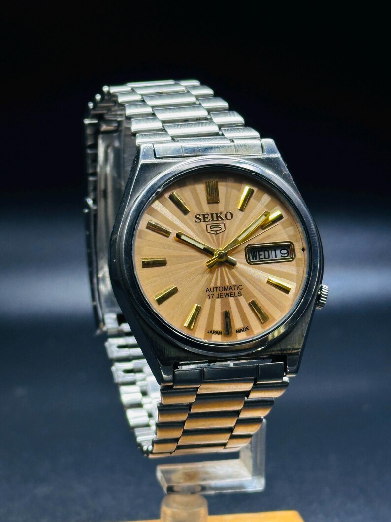 May include: A vintage Seiko automatic watch with a silver stainless steel strap and case. The watch face is light brown with gold hands and markers. The watch displays the day and date. The text "SEIKO" is visible on the watch face.