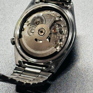 May include: A detailed shot of the back of a silver wristwatch, revealing its internal workings. The metal strap is partially visible, along with the gears and other components of the watch's movement. The watch is set against a grey surface.