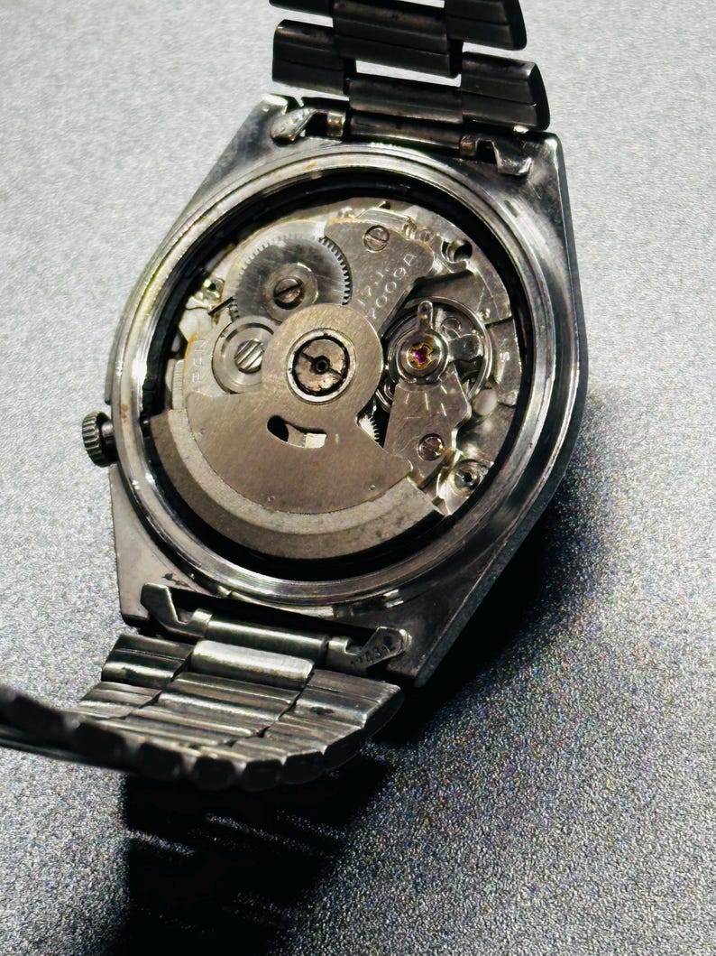May include: A detailed shot of the back of a silver wristwatch, revealing its internal workings. The metal strap is partially visible, along with the gears and other components of the watch's movement. The watch is set against a grey surface.