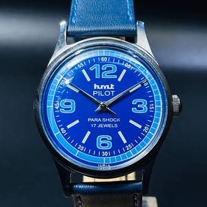 Op de afbeelding: Een blauw en zilveren polshorloge met een blauwe wijzerplaat en witte markeringen. De wijzerplaat van het horloge heeft de woorden "humt PILOT" en "PARA SHOCK 17 JEWELS". Het horloge heeft een bruin leren bandje.