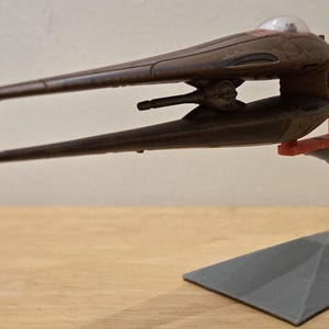 May include: A brown model spaceship with a clear dome and two long, wing-like extensions. The model is mounted on a gray stand with a red support piece. The spaceship has a detailed design and is likely a collectible item.