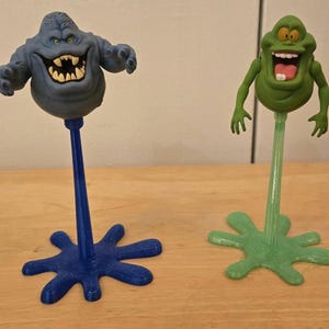 May include: Two novelty figures of cartoon ghosts. One is blue with a wide grin and jagged teeth, mounted on a blue stem and a star-shaped base. The other is green with a large mouth and pink tongue, on a green stem and base.