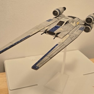 May include: A white and blue model of a Star Wars spaceship with a stand. The spaceship has two engines on the back and two wings that extend out to the sides. The model is weathered and has a worn look.