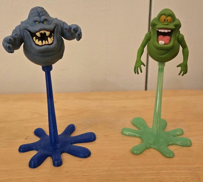 May include: Two novelty figures of cartoon ghosts. One is blue with a wide grin and jagged teeth, mounted on a blue stem and a star-shaped base. The other is green with a large mouth and pink tongue, on a green stem and base.