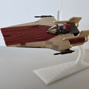 May include: A white and red Star Wars X-wing fighter toy with a pilot figure in the cockpit. The toy is on a white display stand.