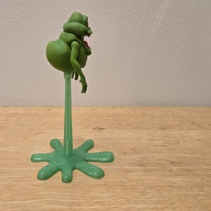 May include: A green, cartoonish figure with a bulbous body and small arms, mounted on a slender green stem and a star-shaped base. The figure has a red mouth and is positioned on a wooden surface.