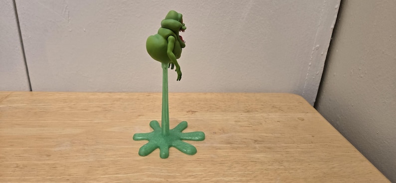 May include: A green, cartoonish figure with a bulbous body and small arms, mounted on a slender green stem and a star-shaped base. The figure has a red mouth and is positioned on a wooden surface.