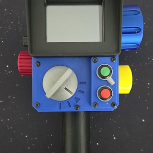 May include: A handheld electronic device featuring a black rectangular screen housing, a blue control panel with a large silver dial, a green button, a red button, and a small toggle switch. The device includes a black handle and side knobs.