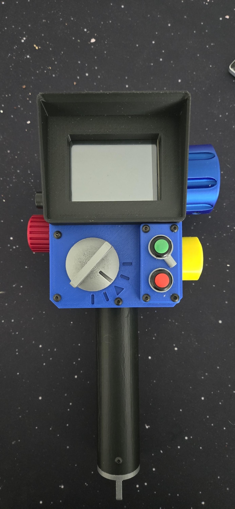 May include: A handheld electronic device featuring a black rectangular screen housing, a blue control panel with a large silver dial, a green button, a red button, and a small toggle switch. The device includes a black handle and side knobs.