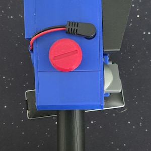 May include: A handheld device featuring a blue rectangular housing with a prominent red circular dial on the top surface. A black handle extends from the base, while a black wire with a connector is attached to the side. A black visor extends from the front.