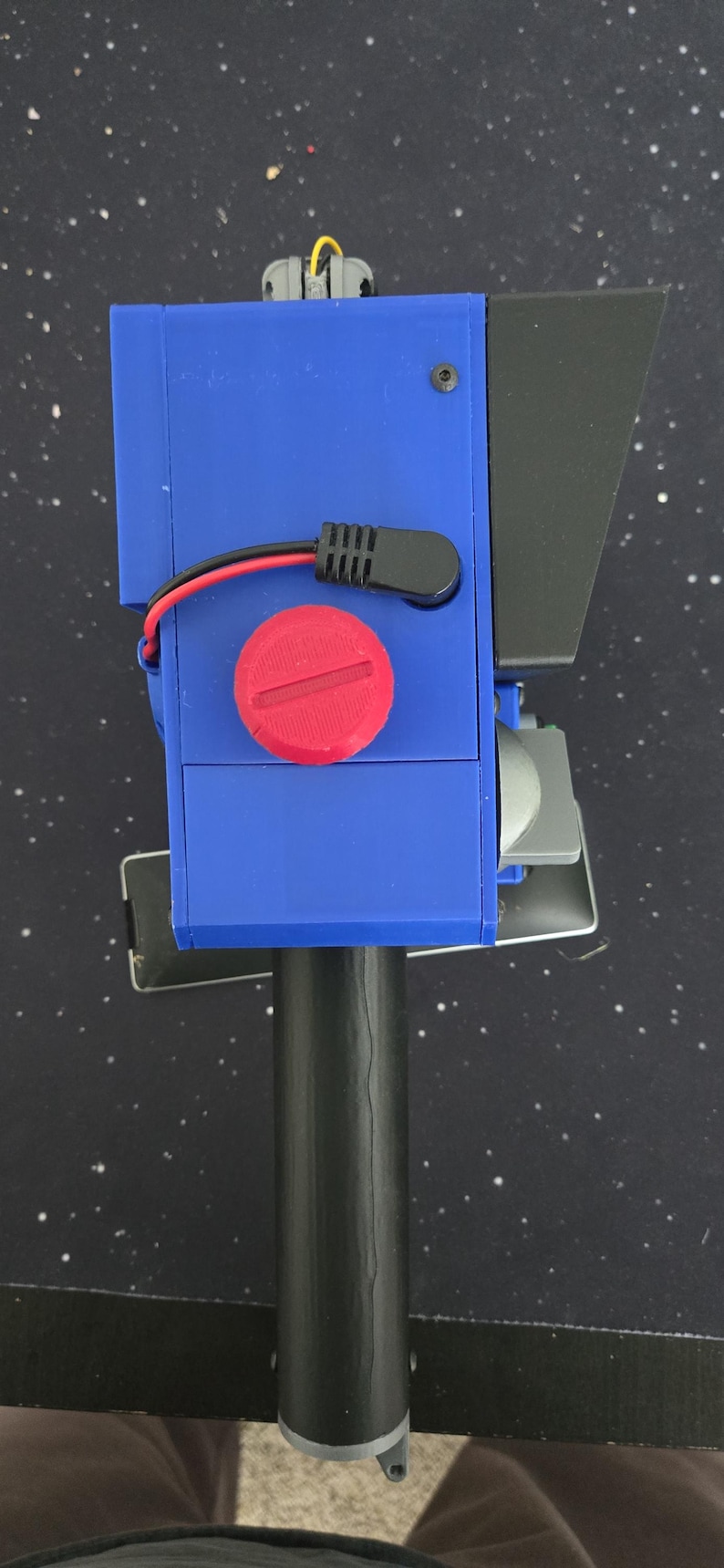 May include: A handheld device featuring a blue rectangular housing with a prominent red circular dial on the top surface. A black handle extends from the base, while a black wire with a connector is attached to the side. A black visor extends from the front.