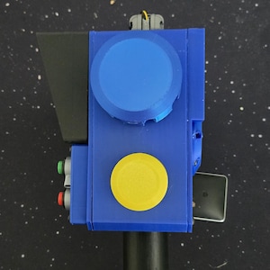 May include: A handheld device featuring a blue rectangular body with a large blue circular dial, a prominent yellow circular button, and two smaller red and green buttons on the side. The device includes a black handle and a black angled visor component.