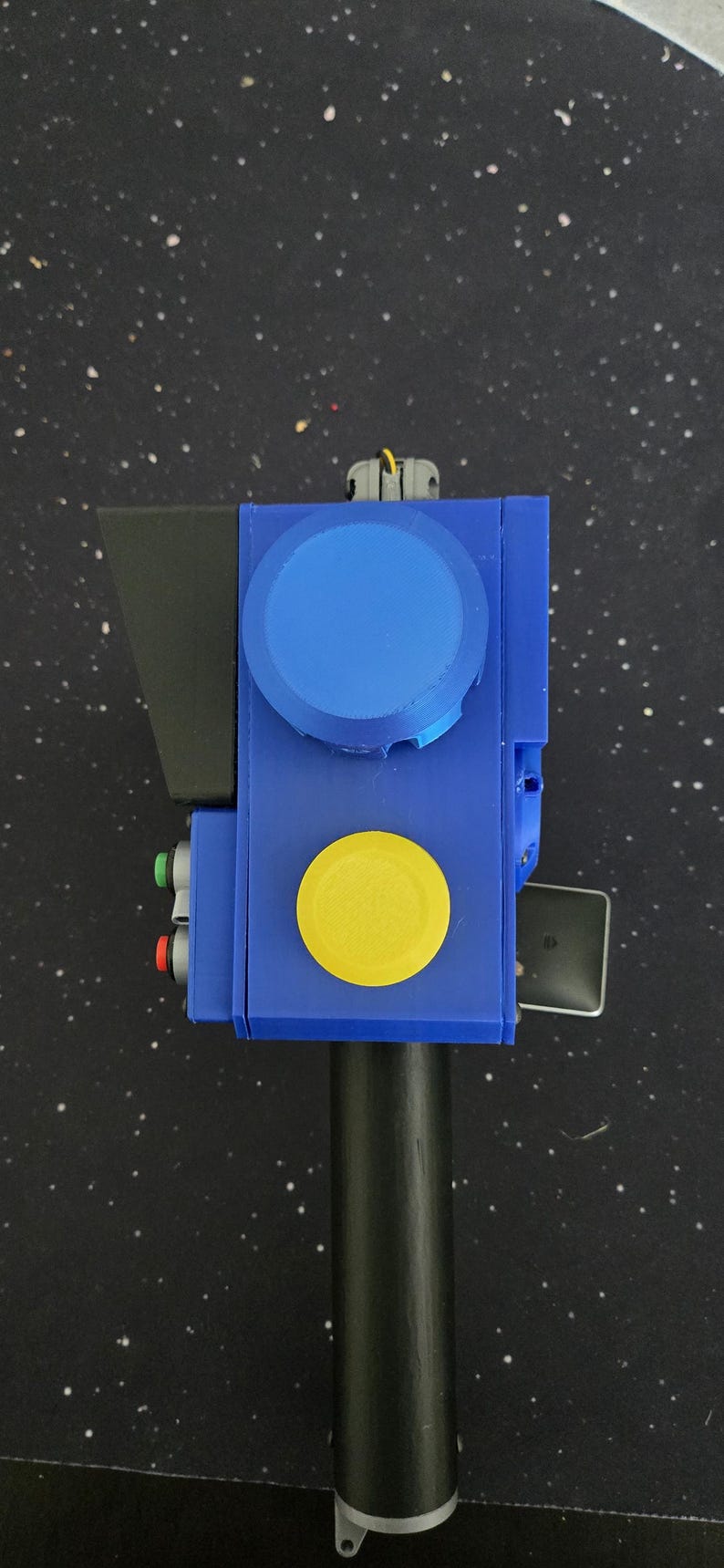 May include: A handheld device featuring a blue rectangular body with a large blue circular dial, a prominent yellow circular button, and two smaller red and green buttons on the side. The device includes a black handle and a black angled visor component.