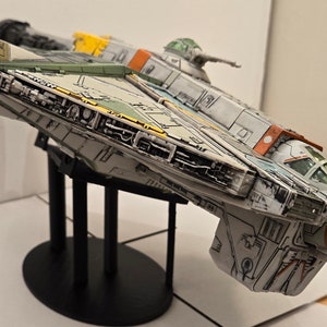 Könnte beinhalten: Ein detailliertes Modell eines weißen, grünen und orangefarbenen Raumschiffs mit einem Cockpit und einem Geschützturm. Das Raumschiff steht auf einem schwarzen Ständer mit runder Basis.