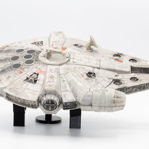 May include: A white and orange model of the Millennium Falcon spaceship from Star Wars. The model is on a black stand and has a weathered look.