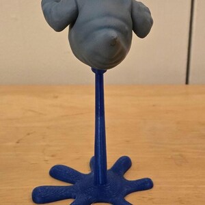 May include: A blue, abstract sculpture of a blob-like figure with outstretched arms, mounted on a slender blue stem and a star-shaped base. The figure is a muted gray color. The sculpture is displayed on a wooden surface.