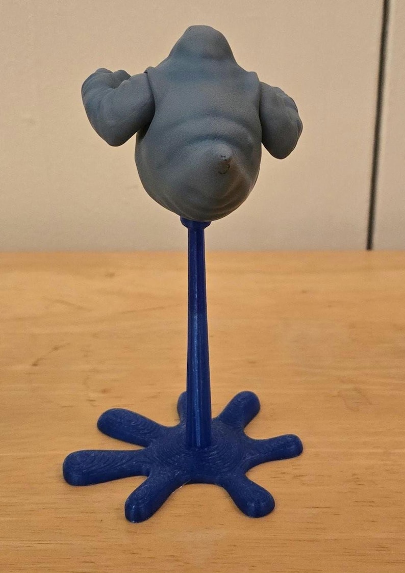 May include: A blue, abstract sculpture of a blob-like figure with outstretched arms, mounted on a slender blue stem and a star-shaped base. The figure is a muted gray color. The sculpture is displayed on a wooden surface.